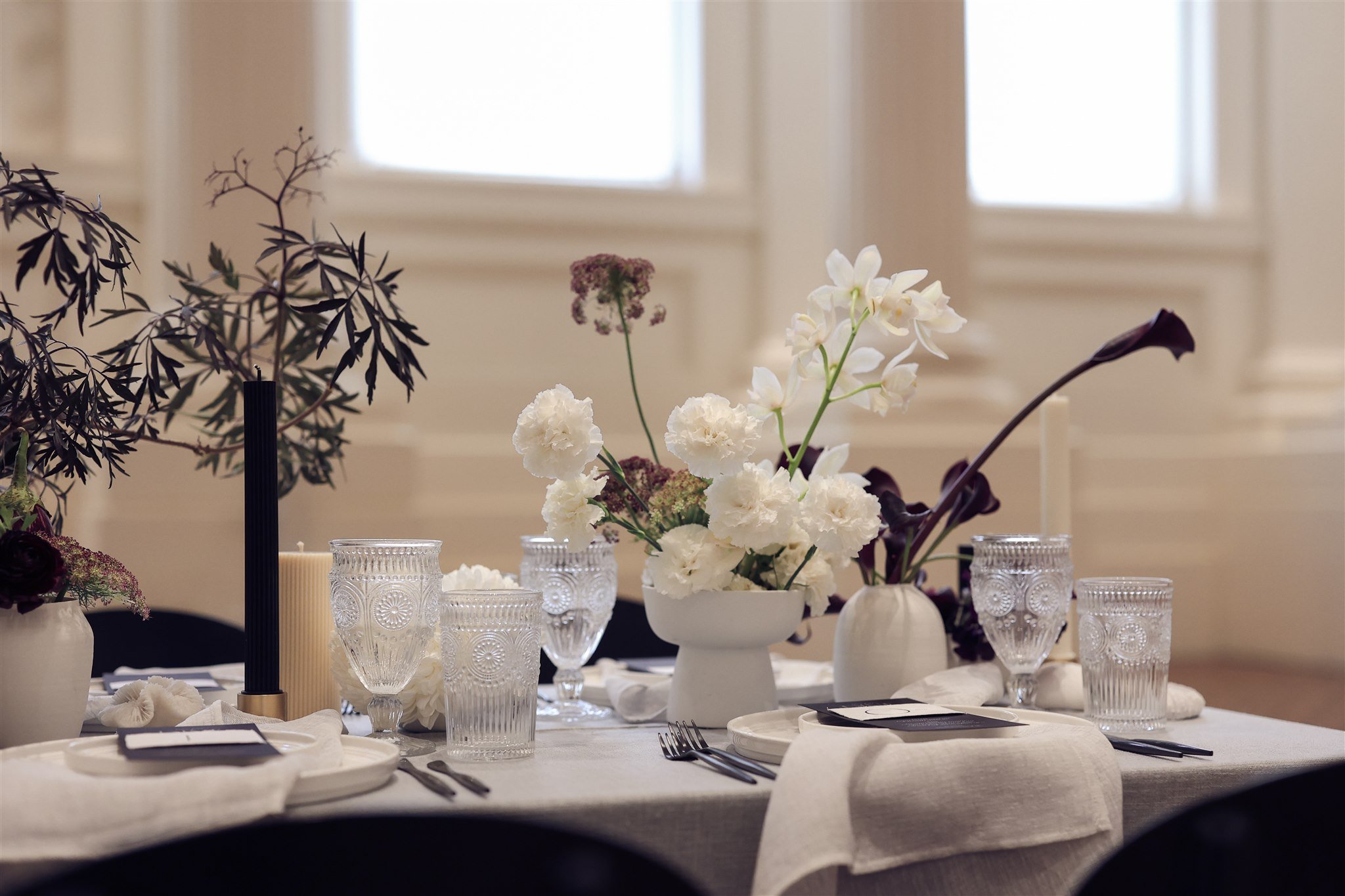 Elegant table setting with white flowers in vases, crystal glasses, dark candles, and white plates on a light tablecloth.