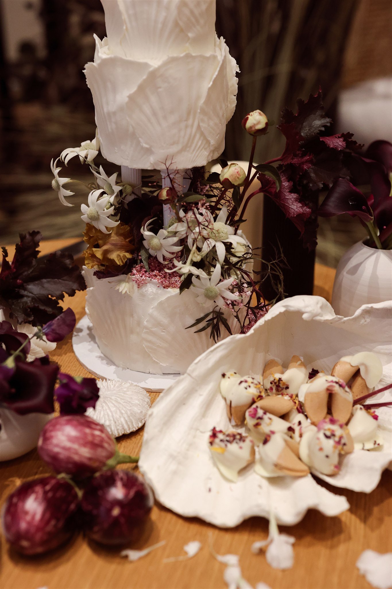 A multi-tiered white cake decorated with flowers and leaves, alongside a large shell-shaped dish filled with decorated cookies on a wooden table.