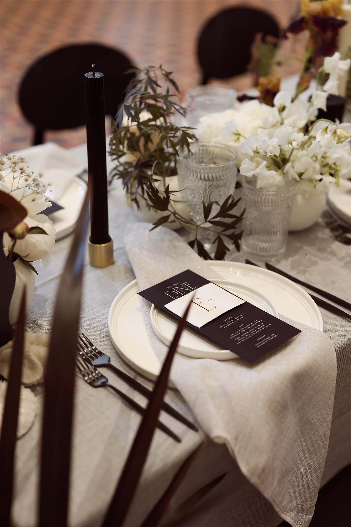 Elegant table setting with white plates, dark cutlery, a black candle, glasses, floral centerpieces, and a printed menu on a beige tablecloth.