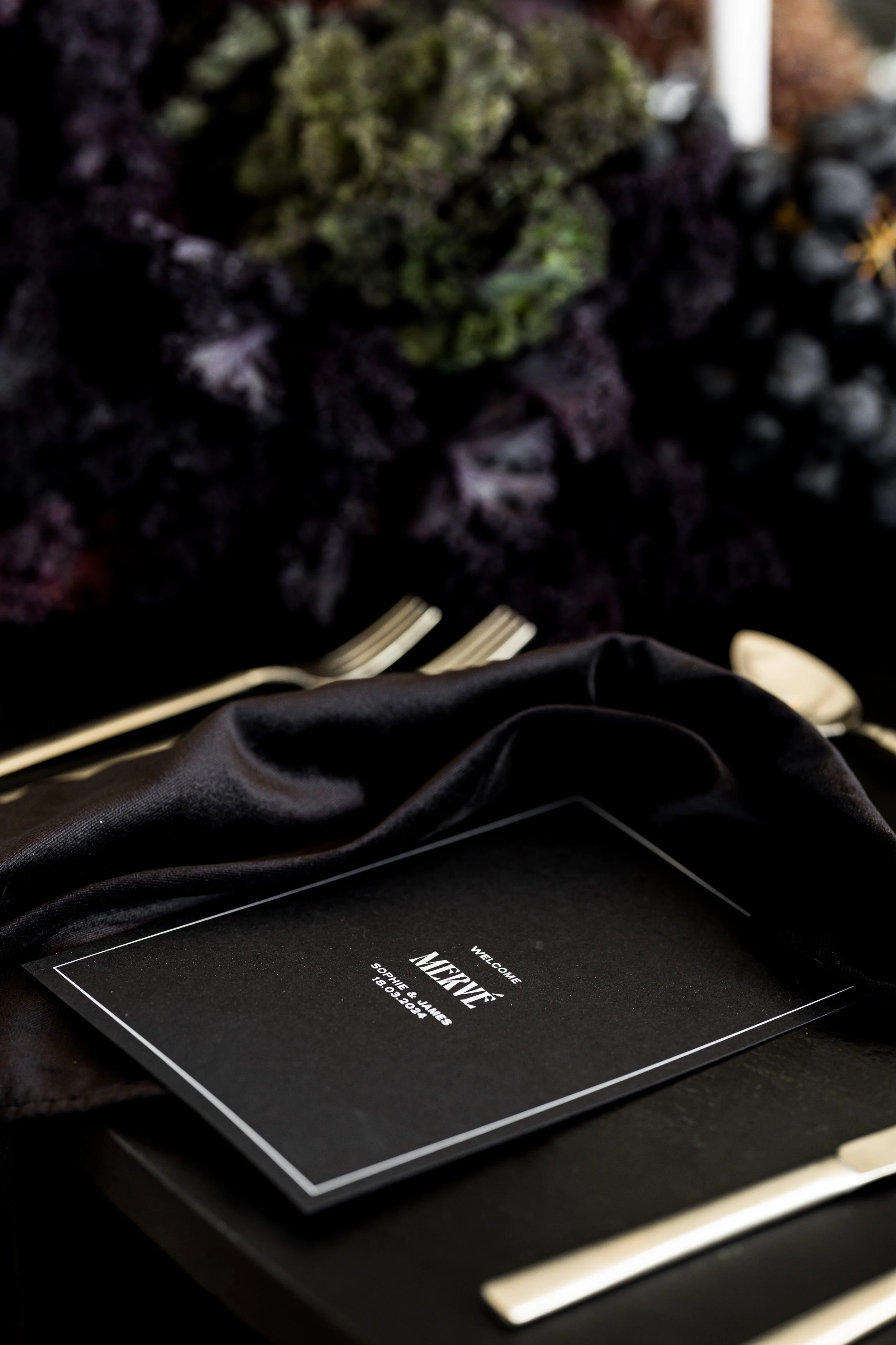 Elegant place setting with a black menu card, silver cutlery, and a black napkin on a dark table, surrounded by dark foliage.