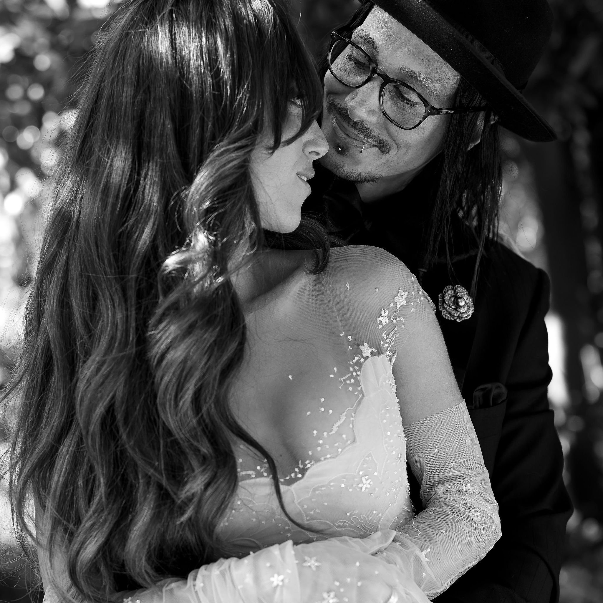 Black and white photo of a couple embracing, with long-haired woman in a detailed dress and man wearing glasses and a hat, looking at each other.