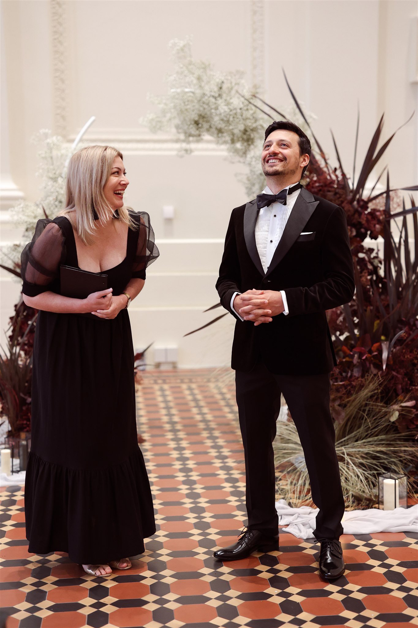 A woman and man dressed in formal black attire, smiling and standing on a patterned tile floor with floral decorations in the background.