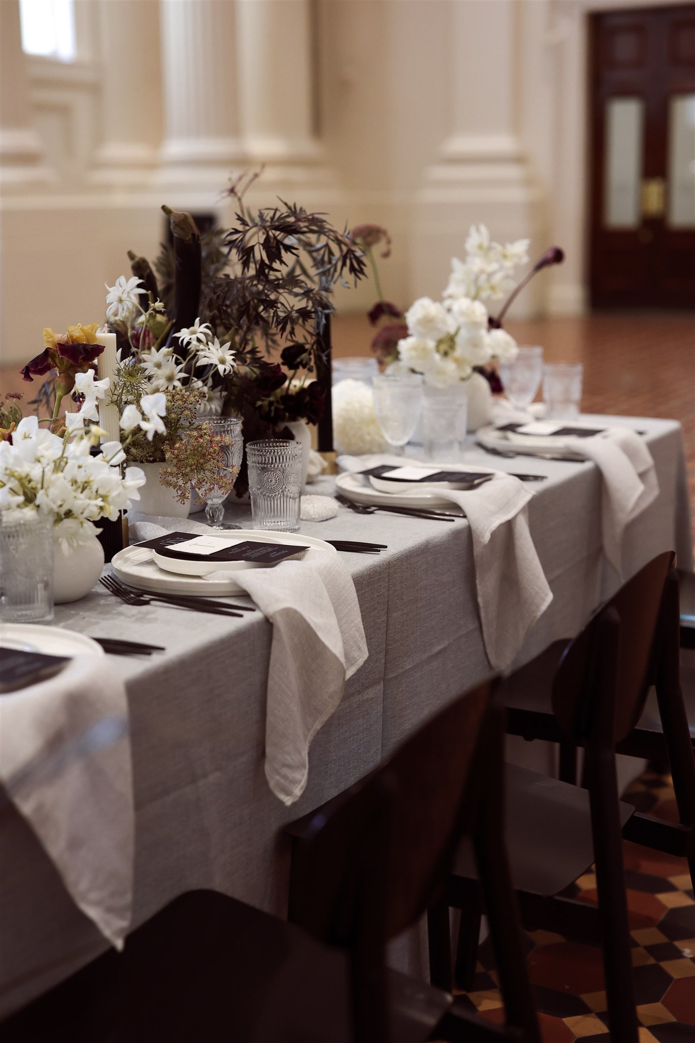 Elegant dining table setup with plates, flatware, and glassware on a grey tablecloth. The table is adorned with white flowers and candles in decorative holders. Neutral napkins are neatly placed, and the setting exudes a refined ambiance.
