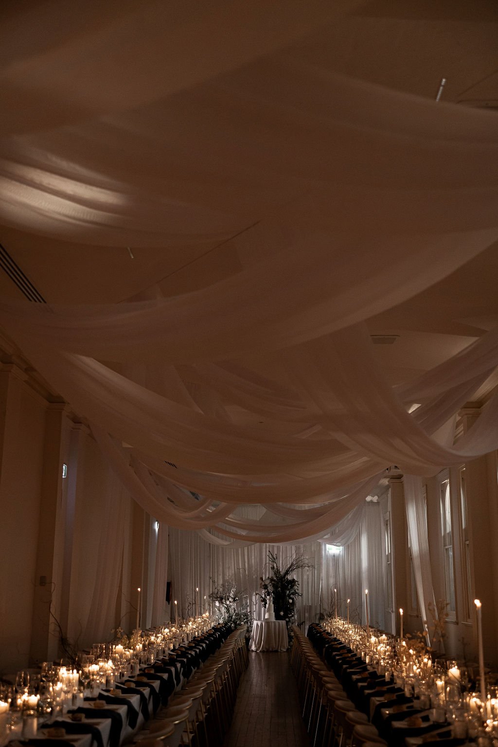 Elegant banquet hall with long tables set for a formal event, featuring candles, white tablecloths, and draped fabric ceiling.