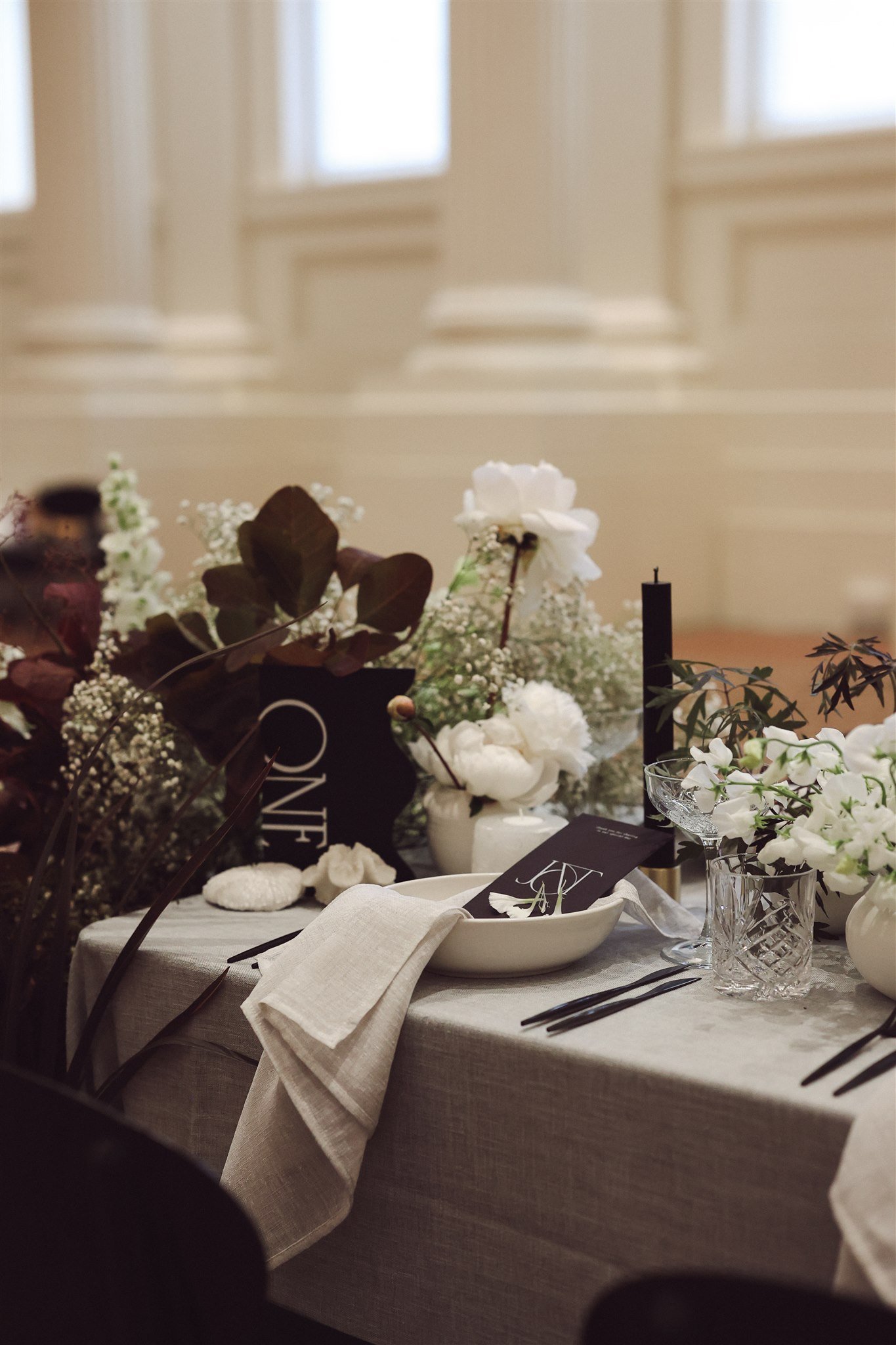 Elegant table setting with white flowers, greenery, a black table number, crystal glassware, and black utensils on a light tablecloth.