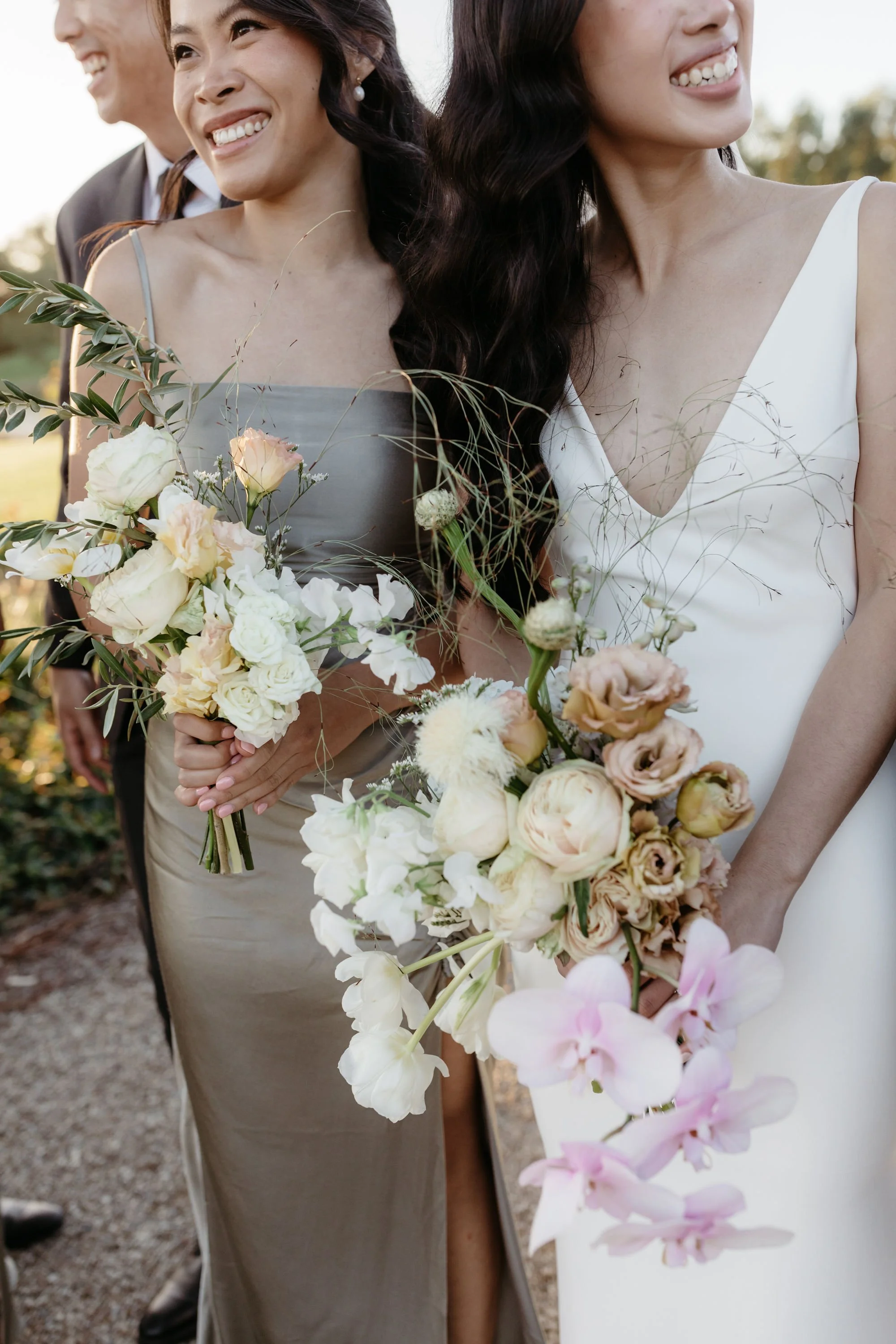 Two women holding bouquets with various flowers, wearing formal dresses, one in white and one in gray.