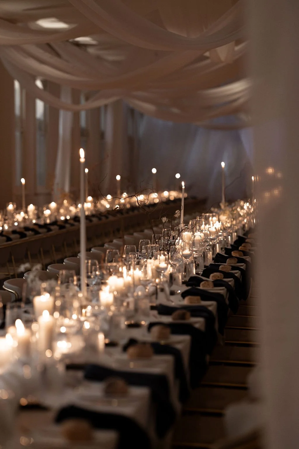 Elegant banquet table set with lit candles, white tablecloths, dark napkins, and decorative branches under draped fabric ceiling.