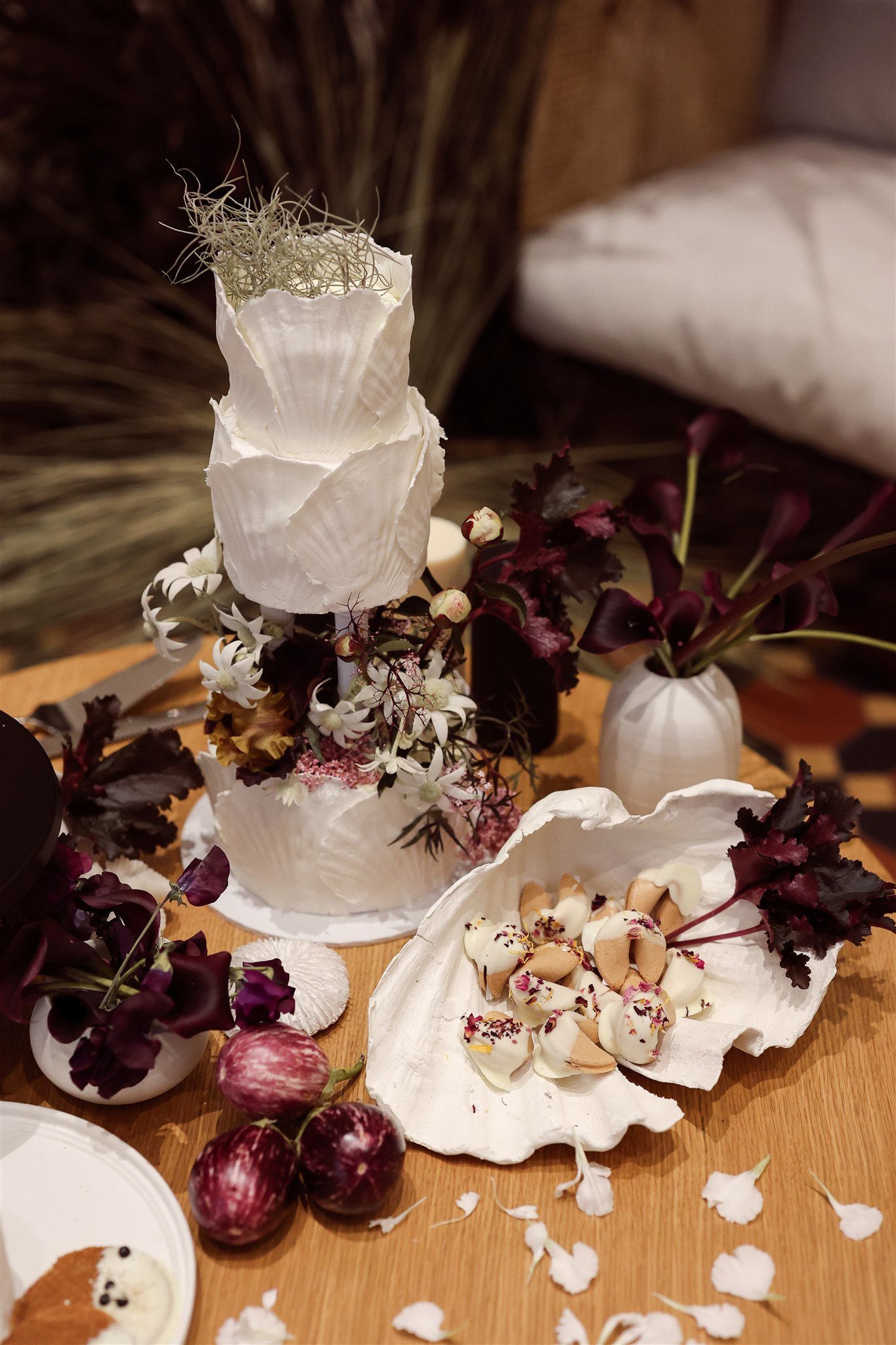 A decorative cake adorned with white seashells and flowers, placed on a wooden table. Nearby are white vases containing purple and dark red flowers, a large white seashell with fortune cookies covered in white chocolate and flower petals, and three p