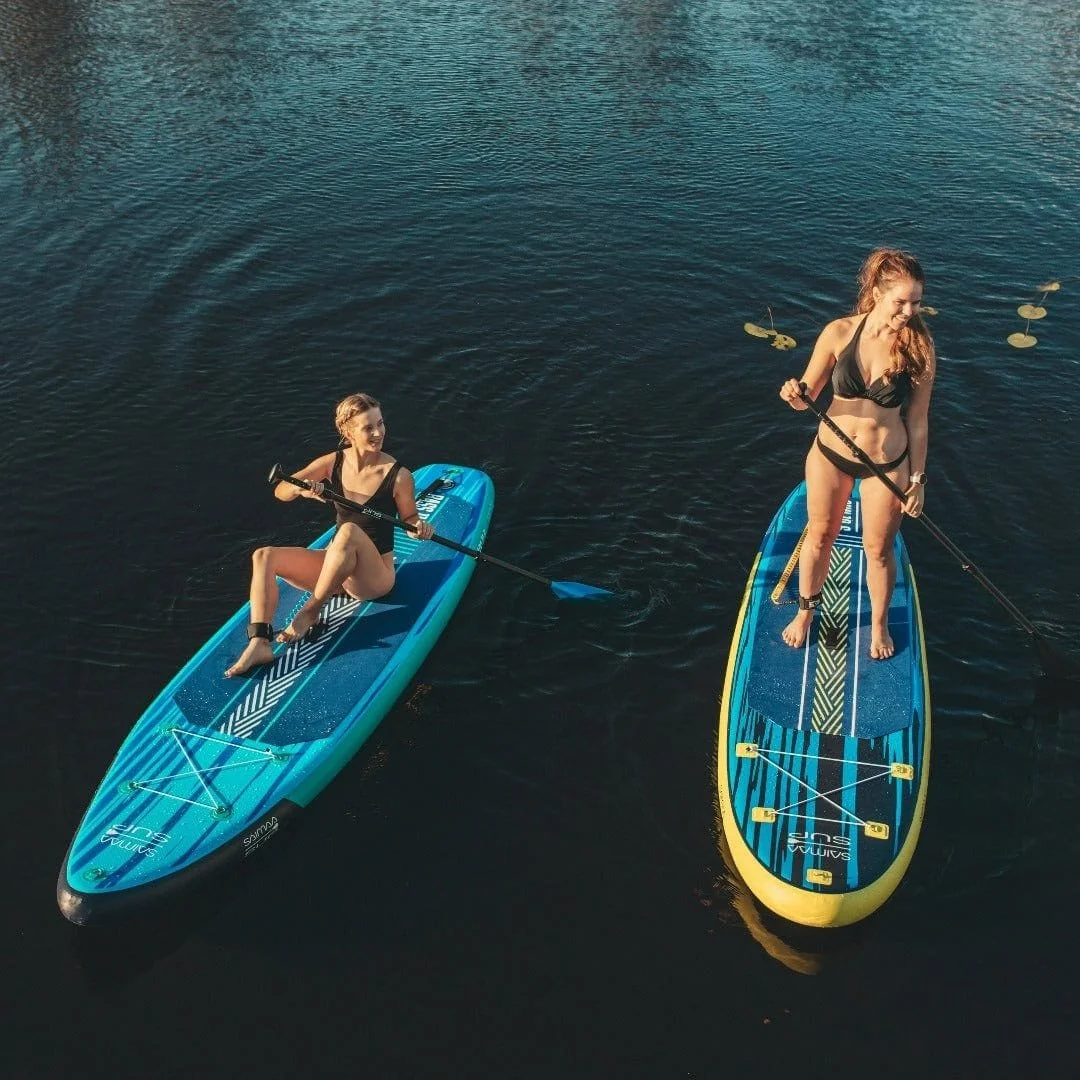 Kaksi naista sup-laudalla vesillä, yksi istuu ja toinen seisoo, molemmat uimapuvuissa.