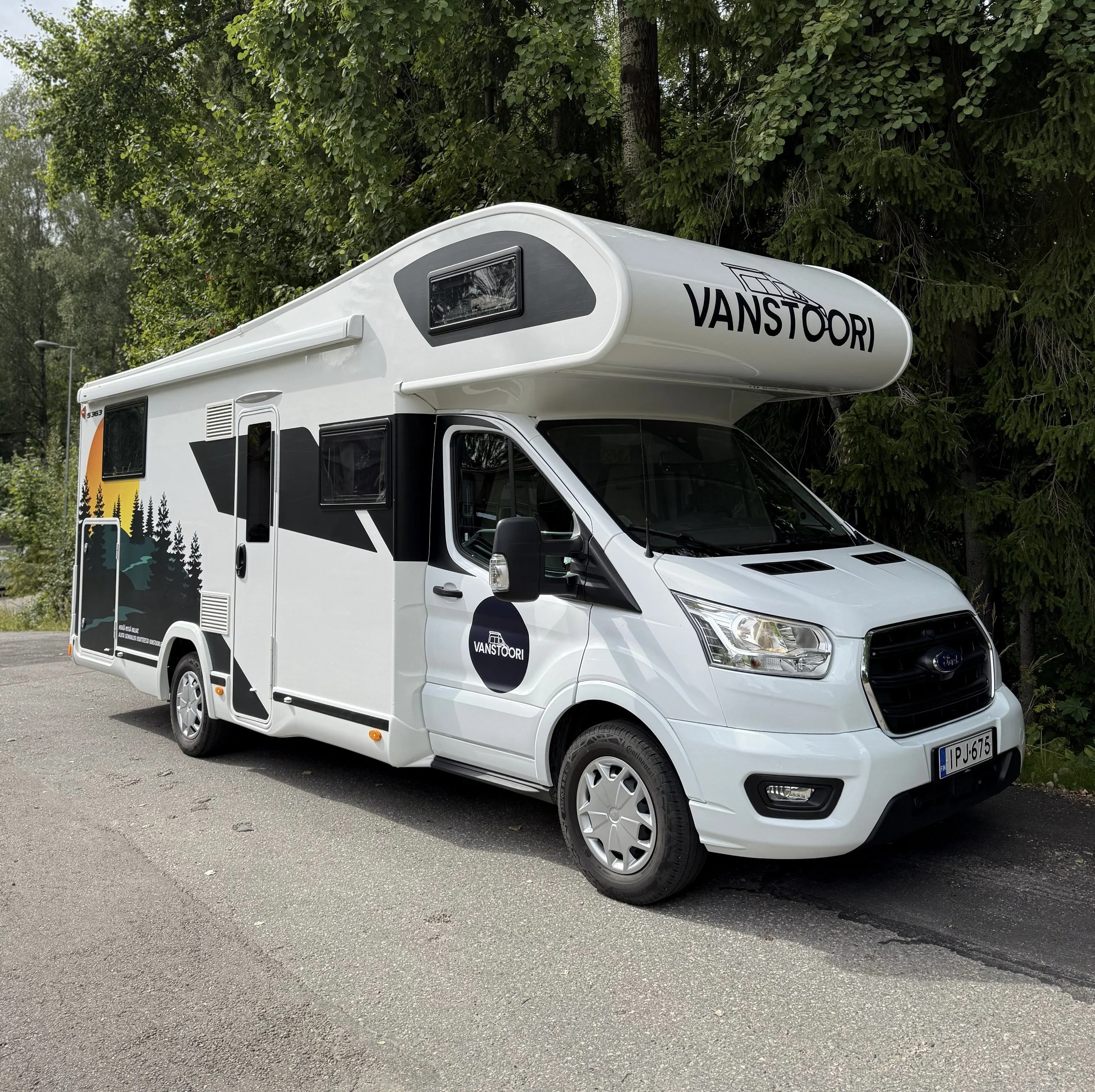 Musta ja valkoinen matkailuauto, jossa "VANSTOORI" teksti yläosassa ja sivulla, pysäköity tielle, taustalla puita.