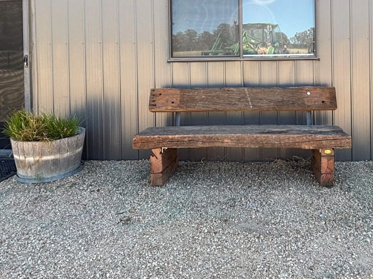 Rustic wooden bench on gravel with metal planter, outside a metal building with window reflecting green farm equipment.