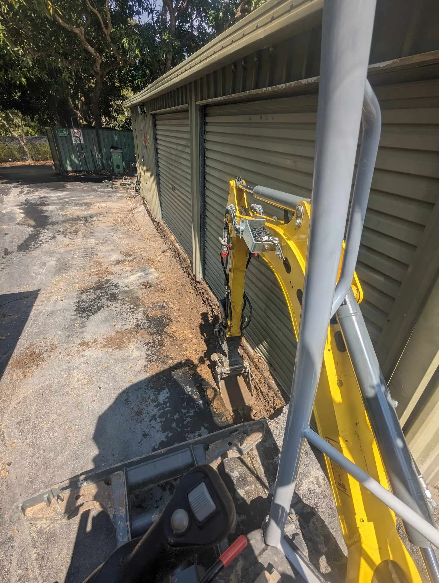 Mini excavator digging along green metal garage with closed doors