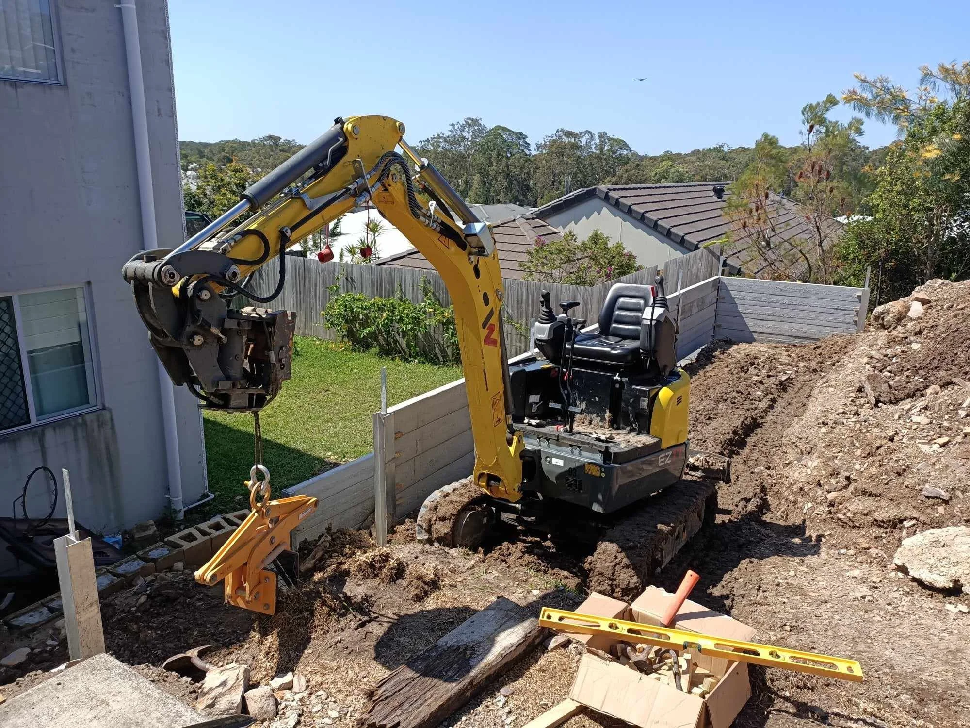 A mini excavator with a grapple attachment at a residential construction site, surrounded by soil and tools, including a spirit level.