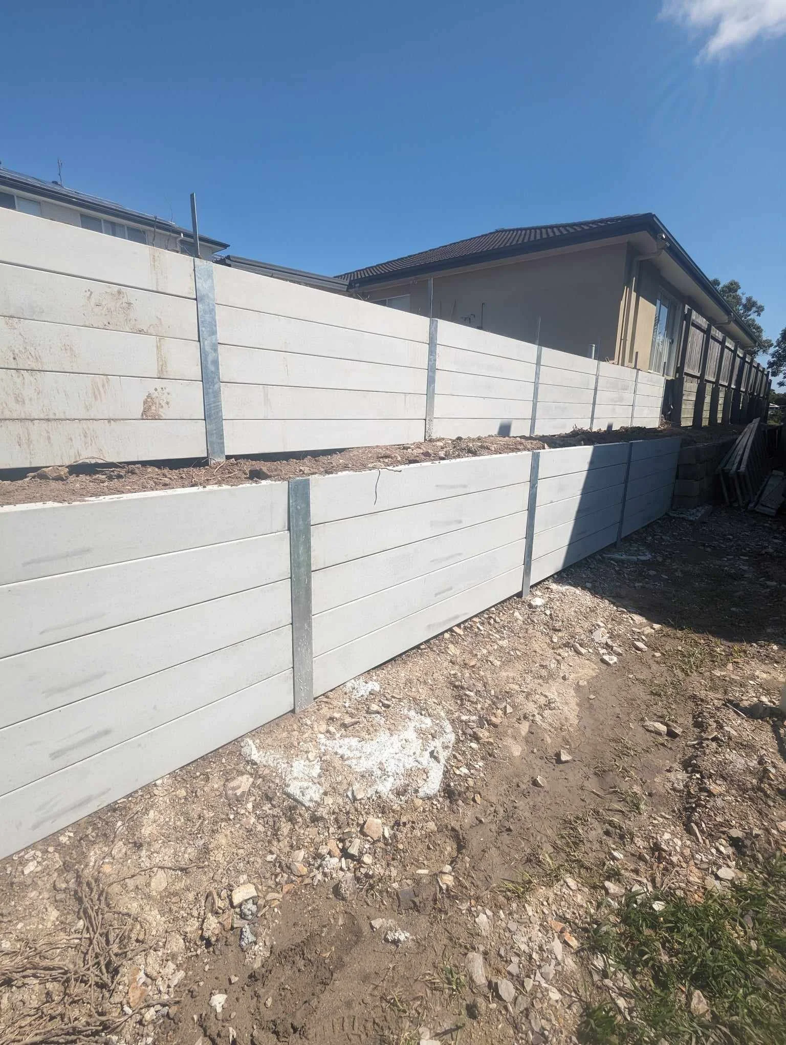 Concrete retaining wall with metal supports next to a house under a blue sky.