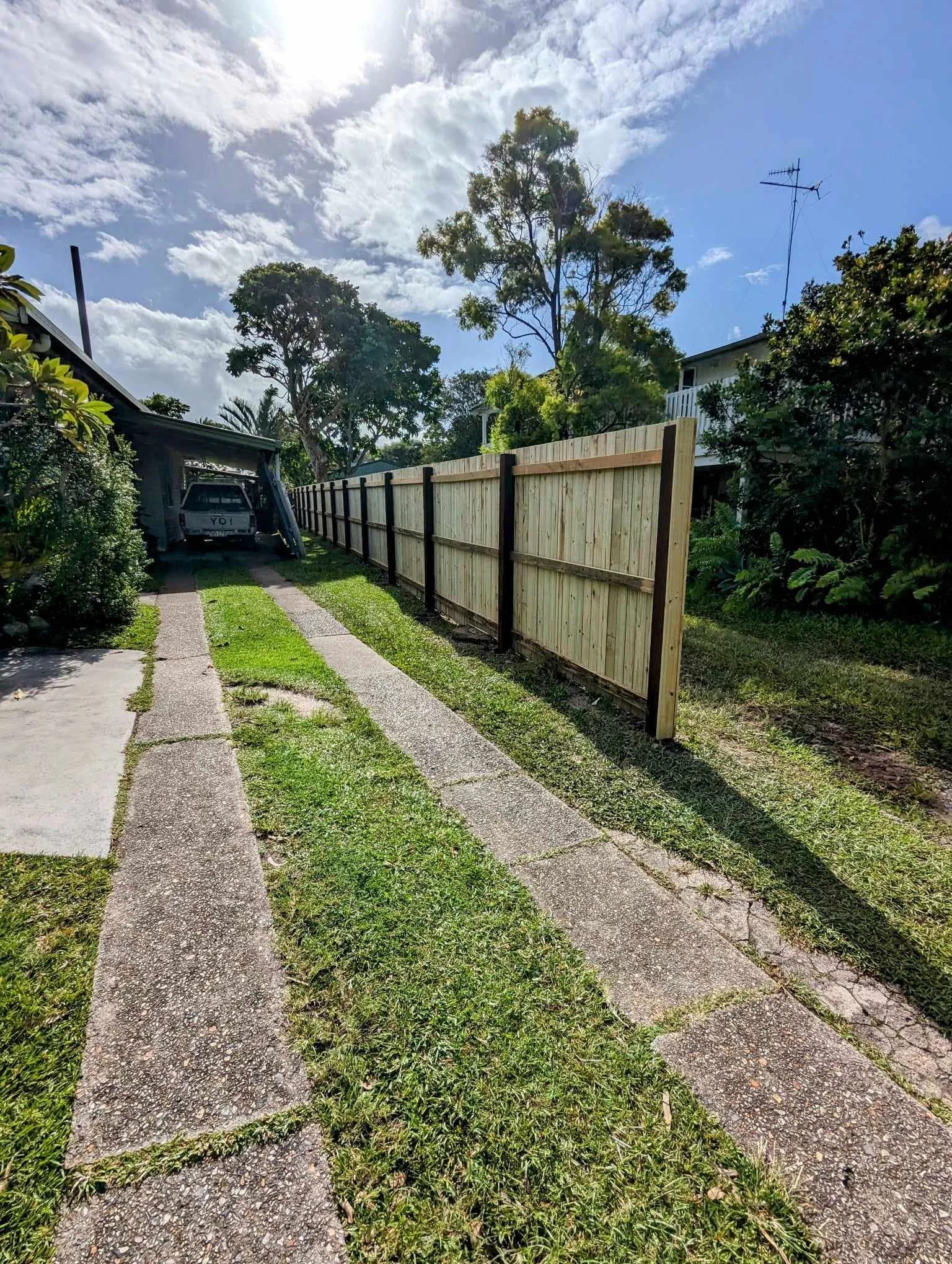 Outdoor scene with a concrete driveway bordered by grass and a wooden fence on the right. A carport contains a vehicle on the left, surrounded by trees and a sunny sky.