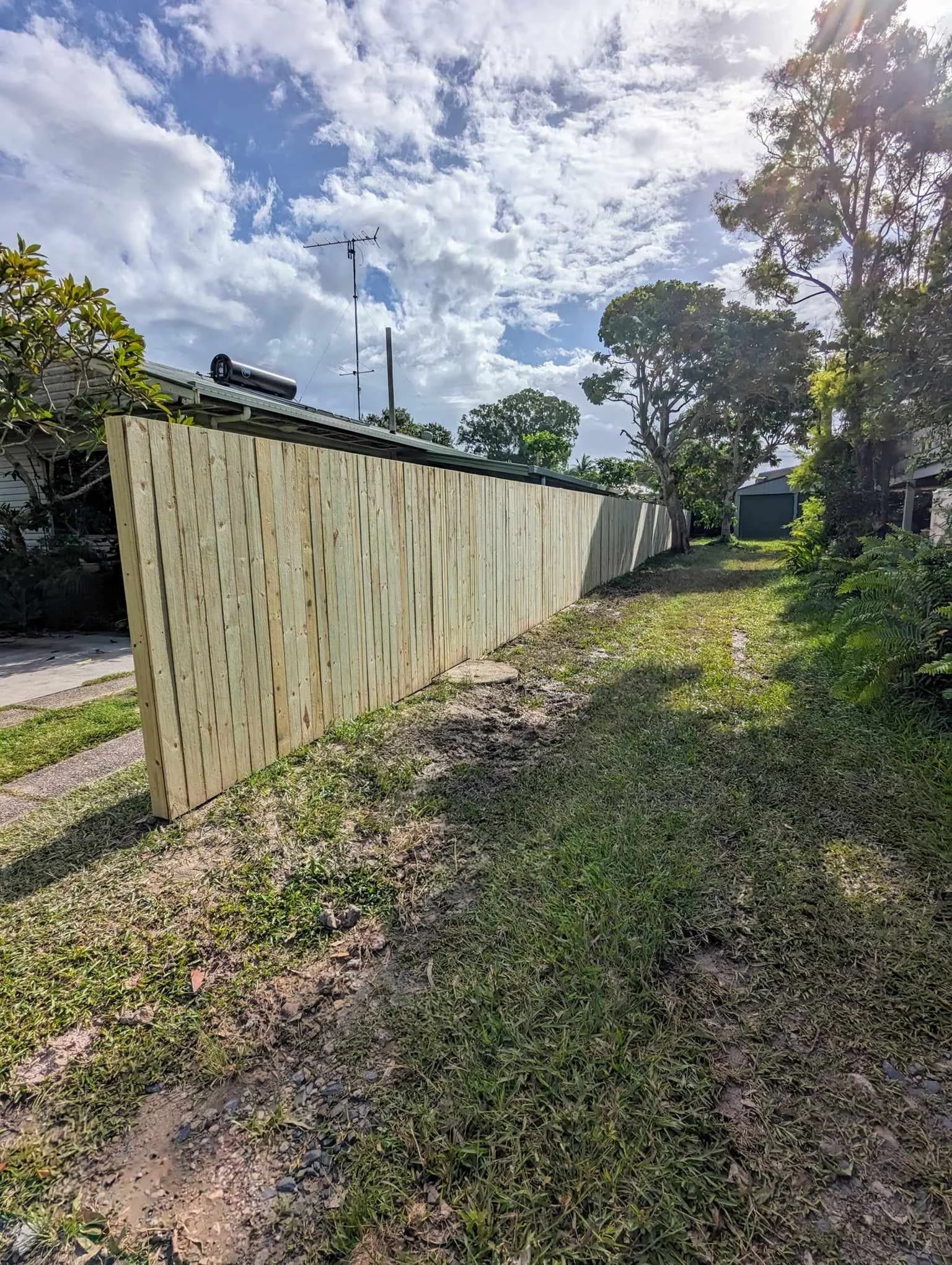 A wooden fence extending along a grassy pathway with trees and a house in the background under a partly cloudy sky.