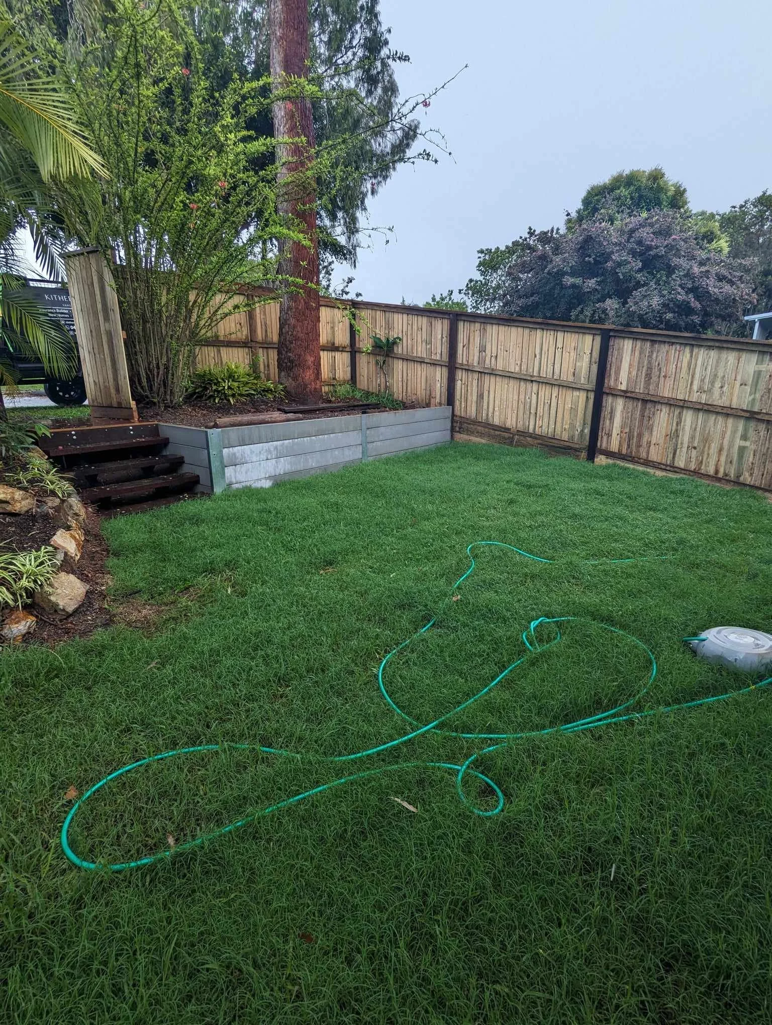 Green grassy backyard with a hose, wooden steps, trees, and a wooden fence.