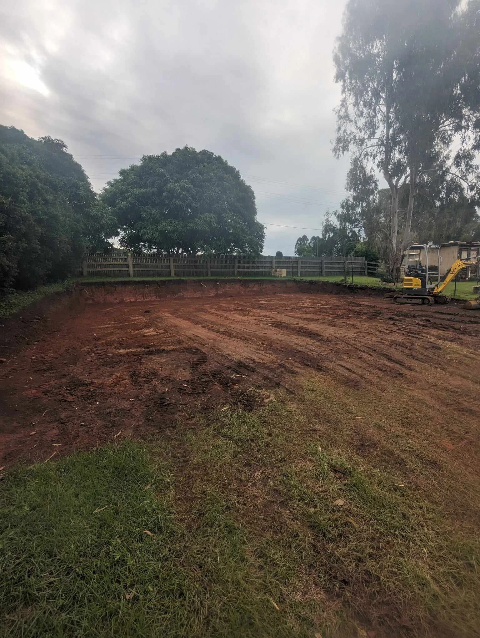 Freshly excavated land next to greenery and trees, with a yellow excavator on the right.