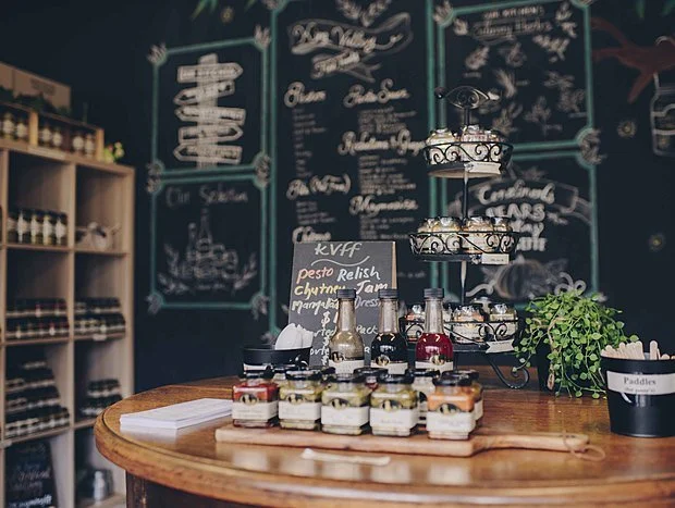 A display table in a shop or cafe with jars of jam, bottles of syrup, and a small potted plant. Chalkboard menus are visible in the background.