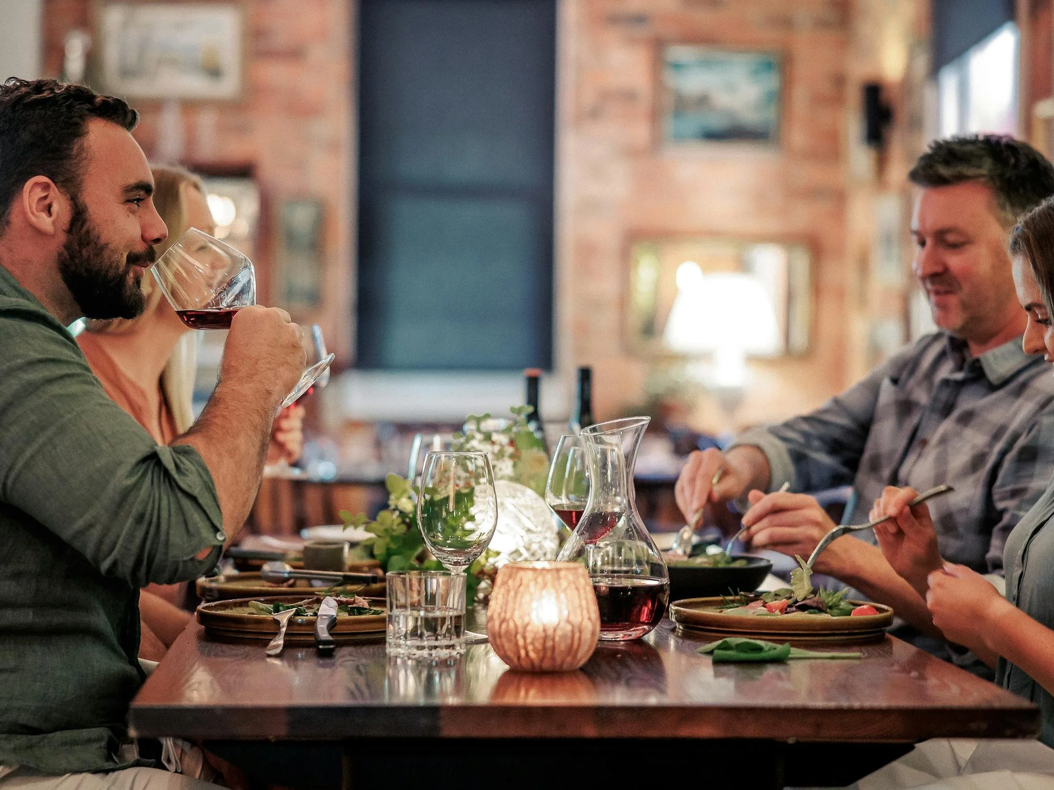 Group of people enjoying a meal at a dinner table in a cozy, warmly lit restaurant, with wine glasses, plates, and a candle.