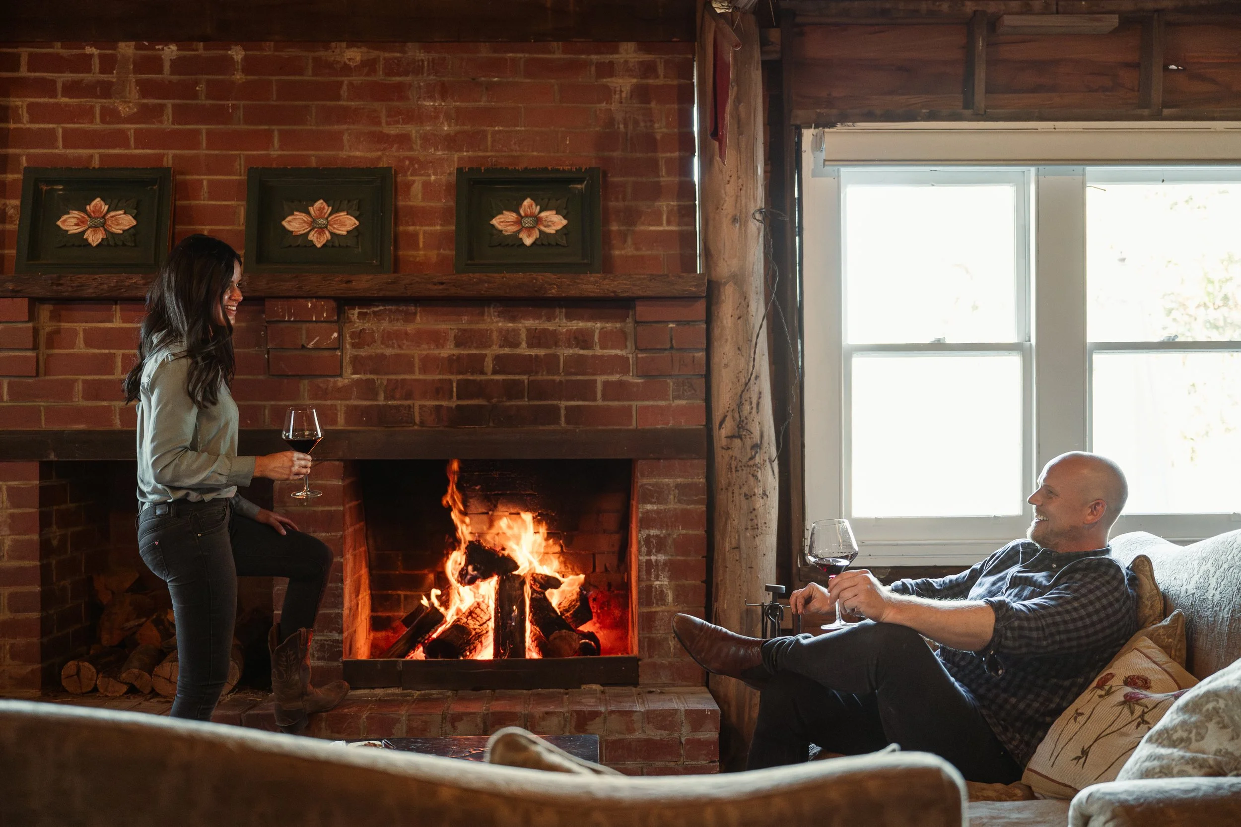 A woman and a man enjoying drinks by a fireplace in a cozy, rustic living room with brick walls and large windows.