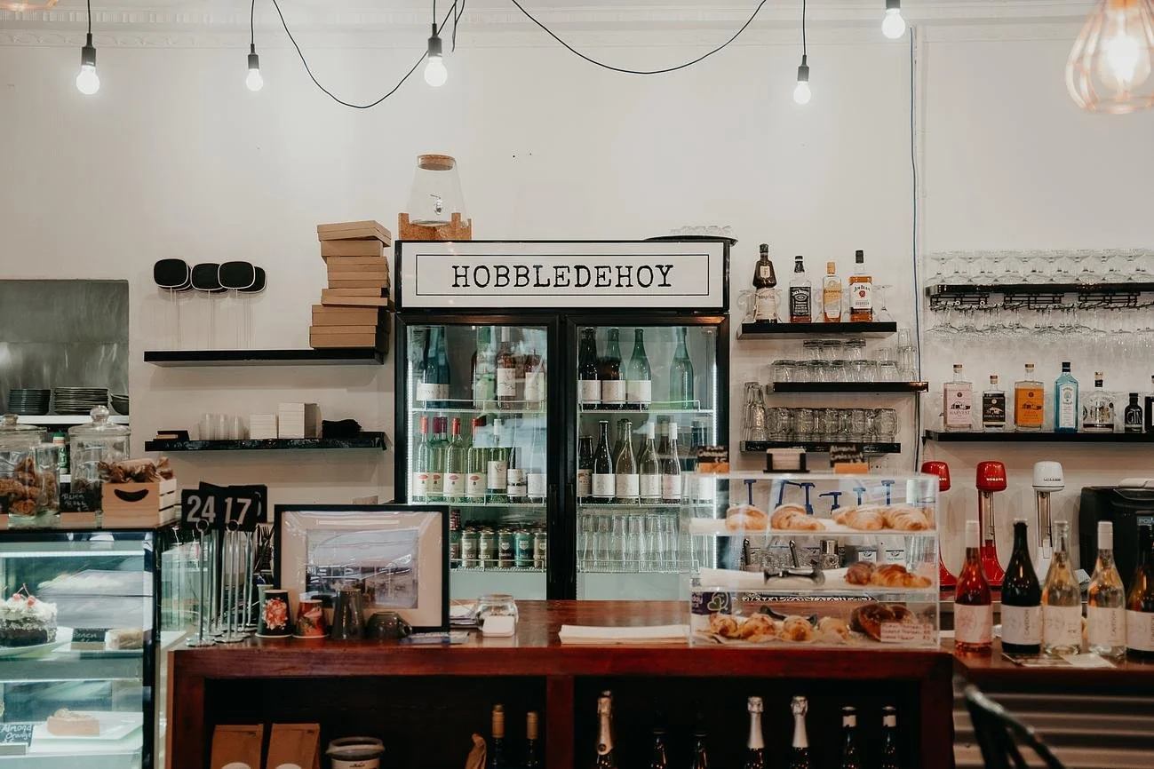 Coffee bar with a glass display case of baked goods, bottles of wine and liquor, glassware, and shelves with books, glasses, and bottles. Sign reads 'HOBBLEDHOY'.