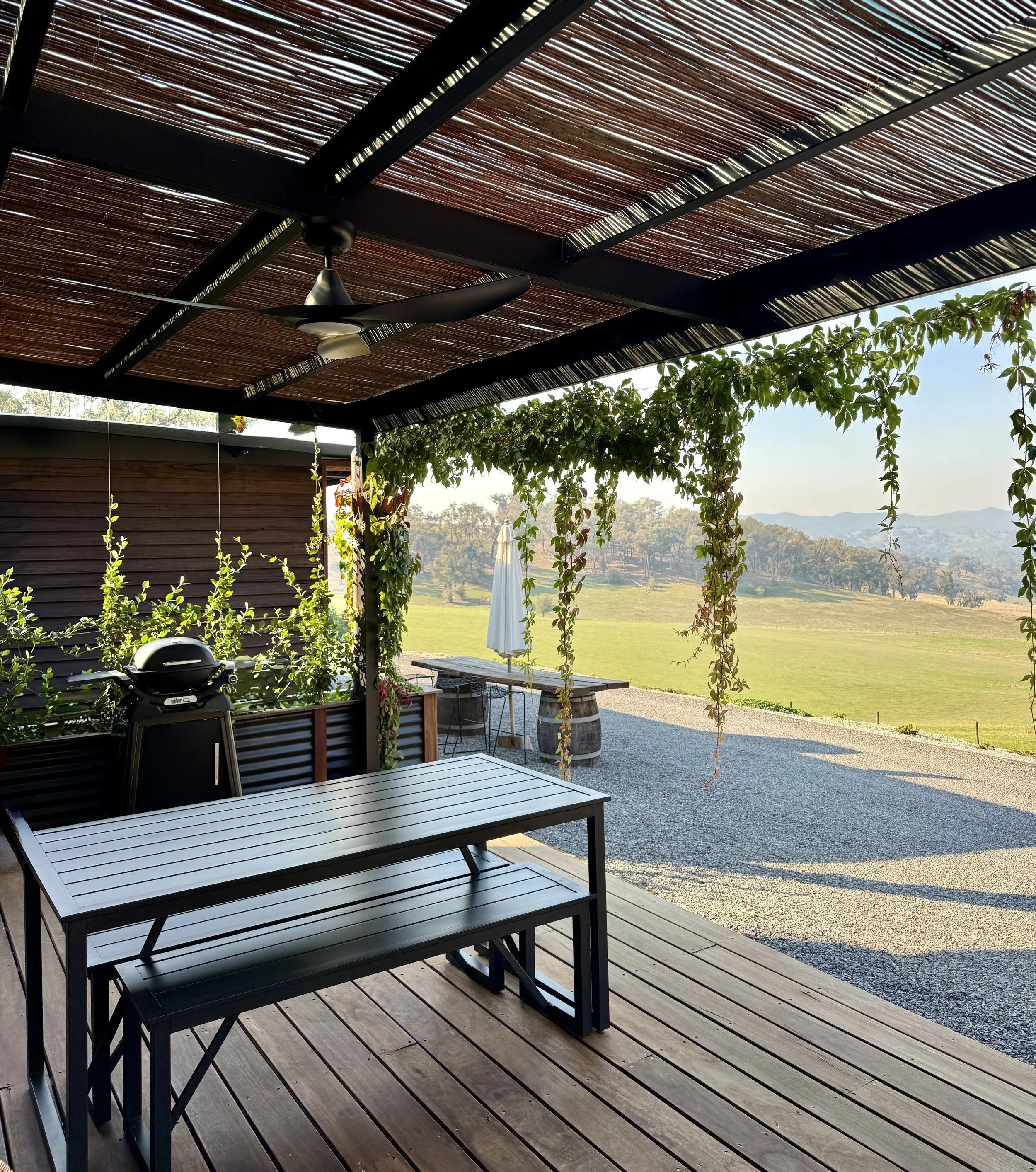 Outdoor patio with a table and benches, a ceiling fan, a grill, and a view of green fields and rolling hills in the distance.