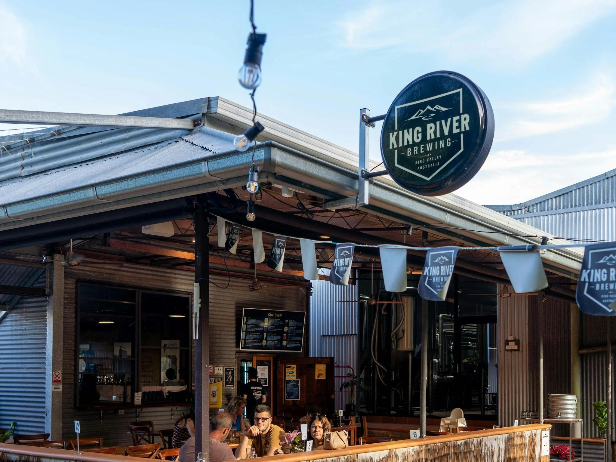 Outdoor seating area of King River Brewing with customers sitting at tables under the sign, metal building, hanging string lights, and menu on display.