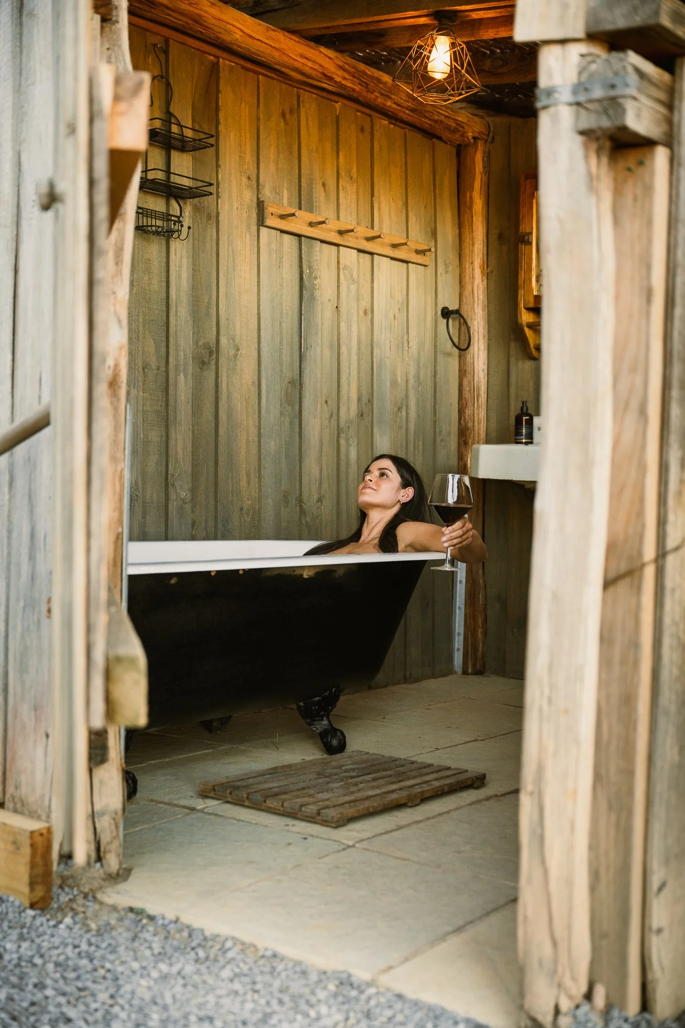 A woman relaxing in a vintage clawfoot bathtub with black exterior, holding a glass of red wine, in a rustic wooden bathroom.