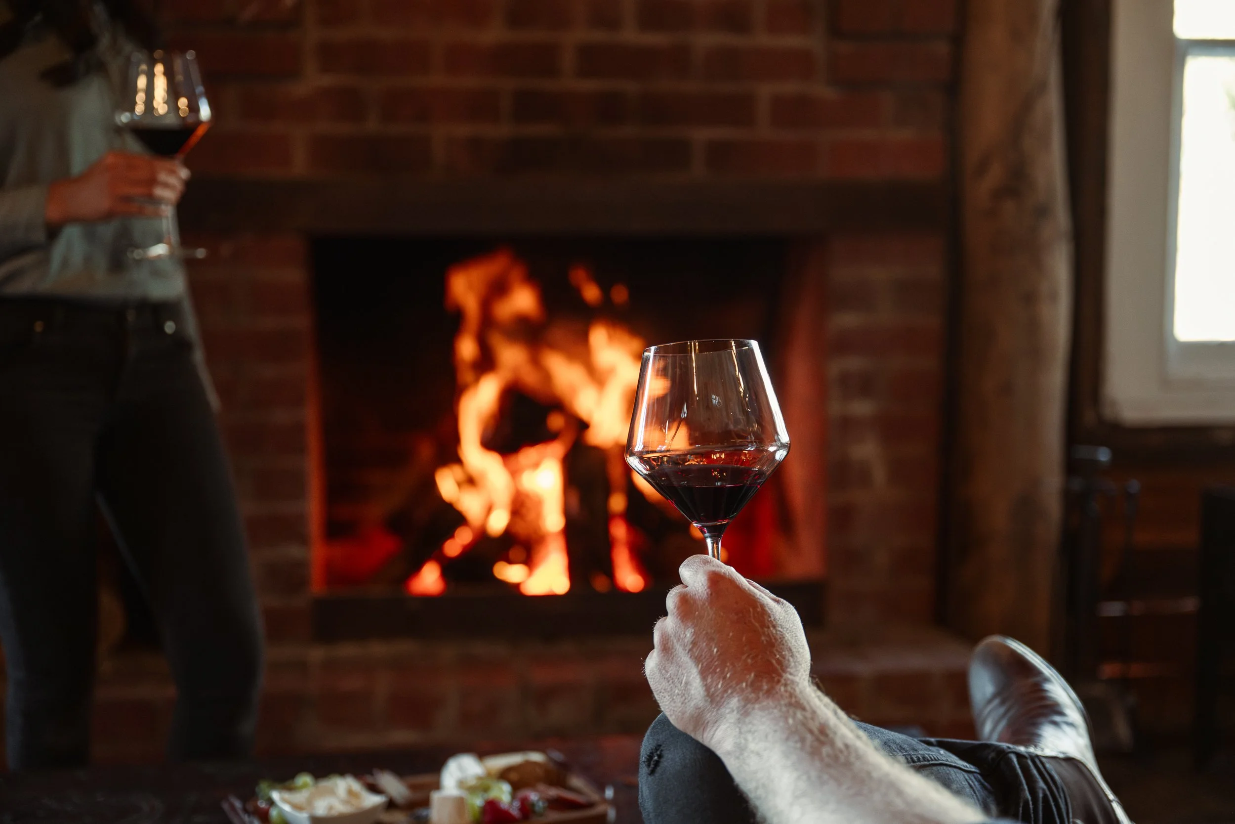 Person relaxing with a glass of red wine by a cozy fireplace, while another person stands holding a glass of wine in the background.