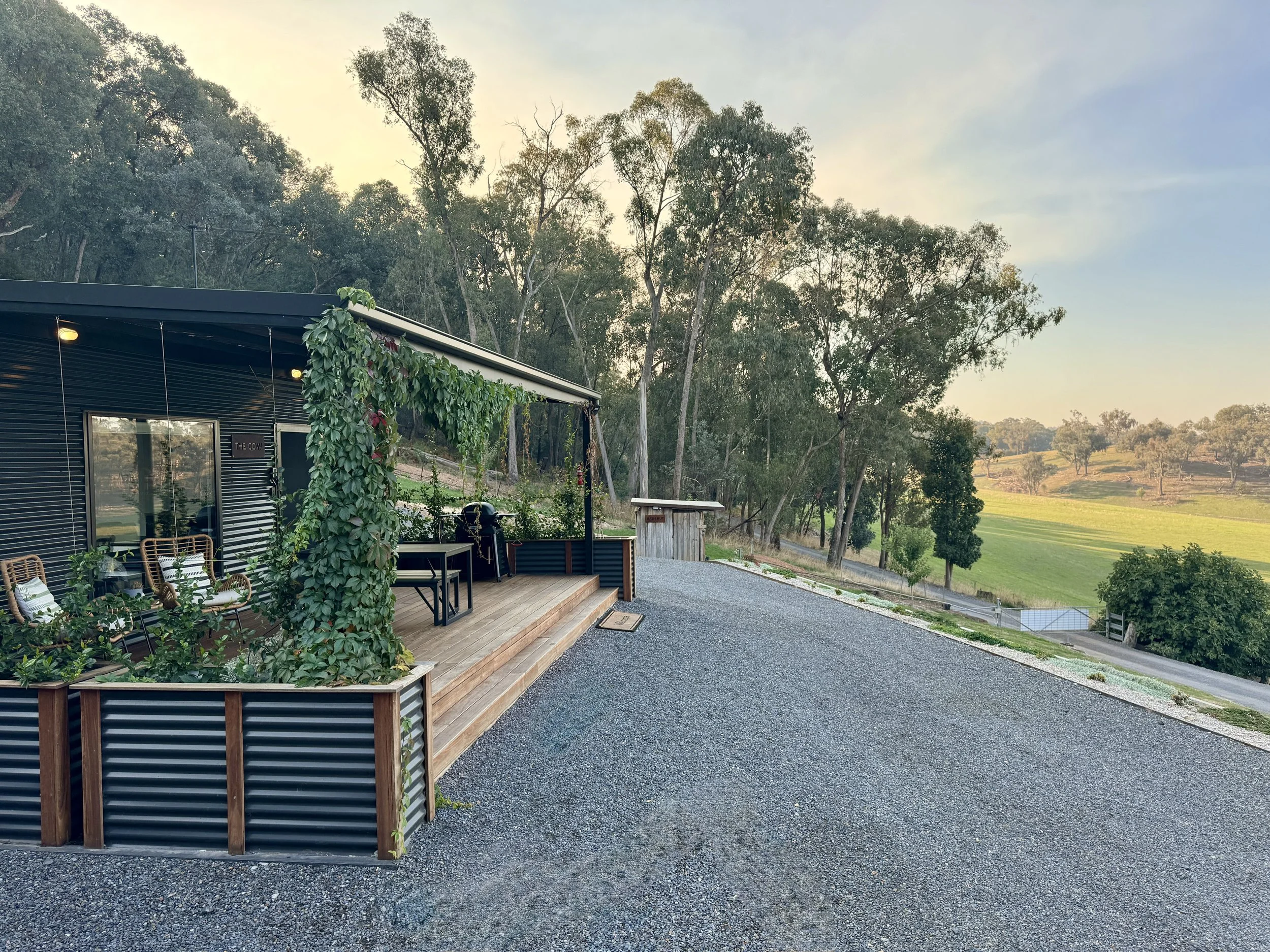 House with a wooden deck, black metal siding, and green vines, set in a rural landscape with trees and open fields under a partly cloudy sky.