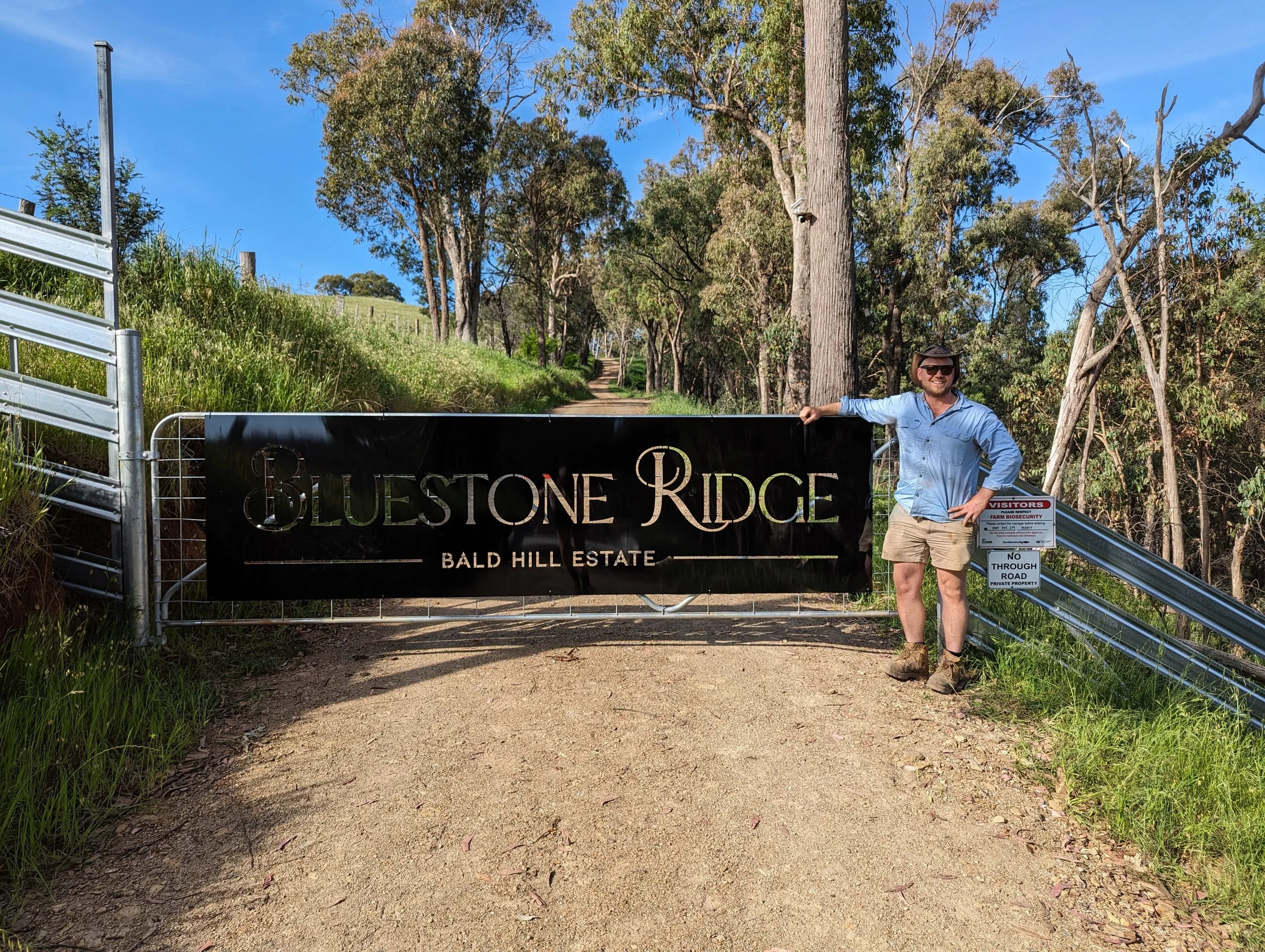 A man standing next to a black gate with a sign that reads 'Bluestone Ridge Bald Hill Estate' in a rural area with trees and a dirt road. The man is wearing sunglasses, a blue shirt, shorts, and hiking boots.