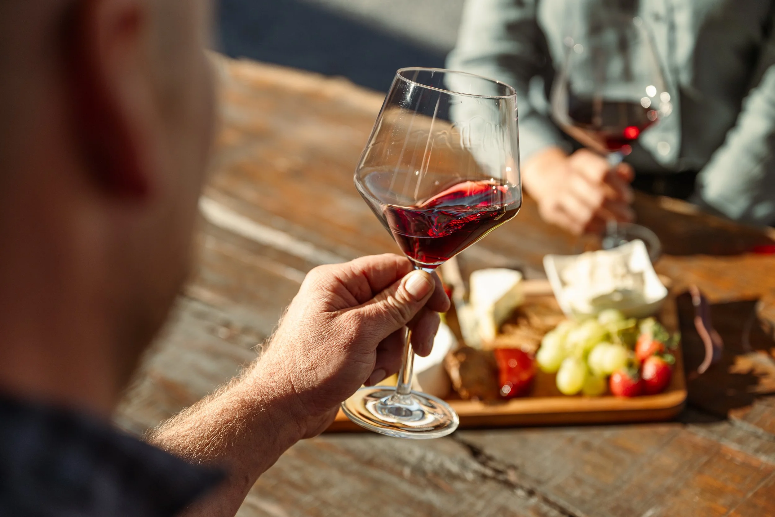 A person holding a glass of red wine with a cheese and fruit platter on the table in front of them, during a social gathering.