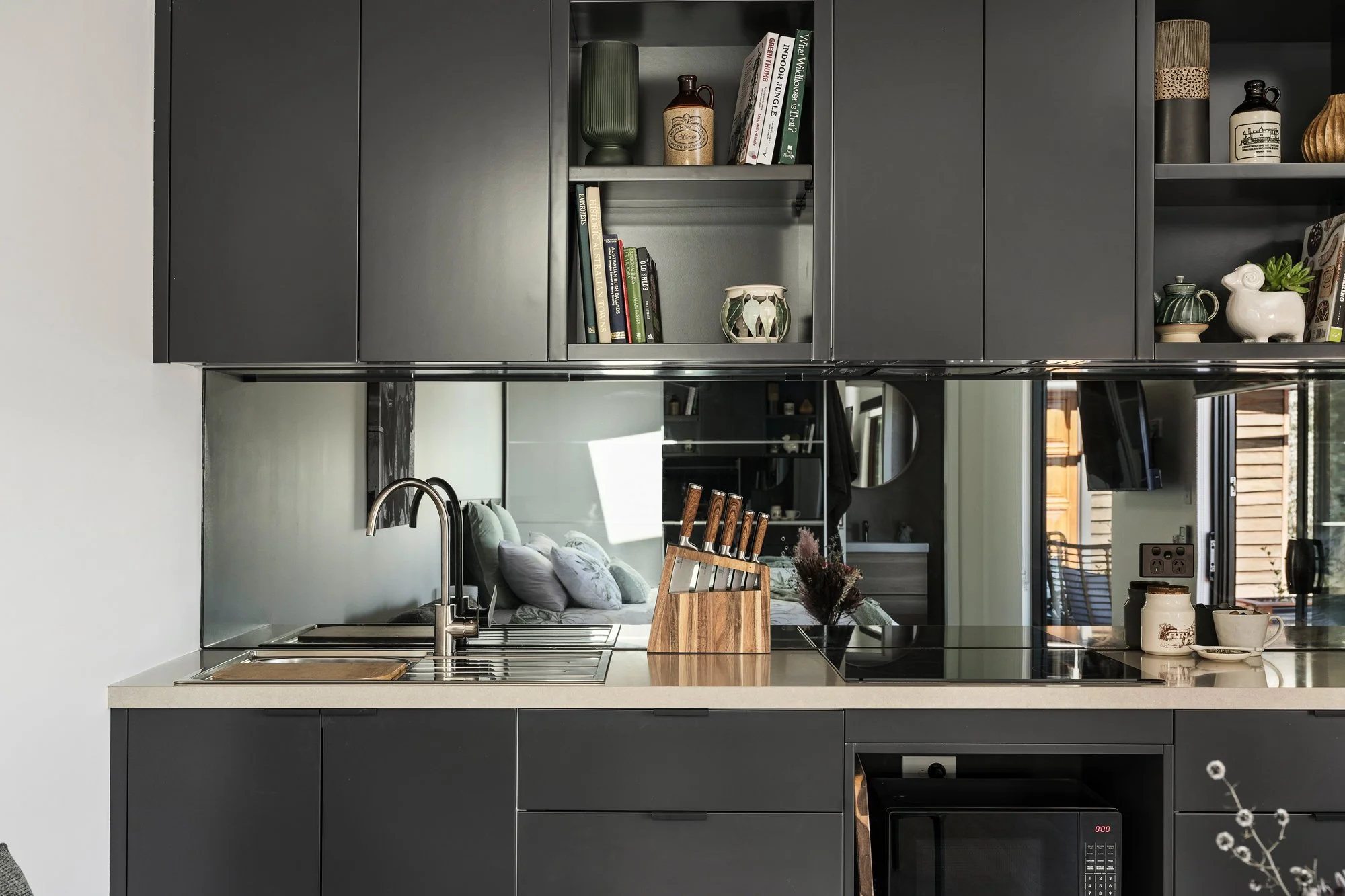 Modern kitchen with black cabinets, beige countertop, and a mirror wall reflecting a living area with a sofa and pillows. The kitchen features a sink, a knife block, and some cups and jars on the countertop.