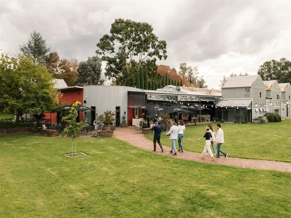 People walking towards an outdoor event space at a modern mixed-use building with patio seating and string lights on a cloudy day, with trees in the background.