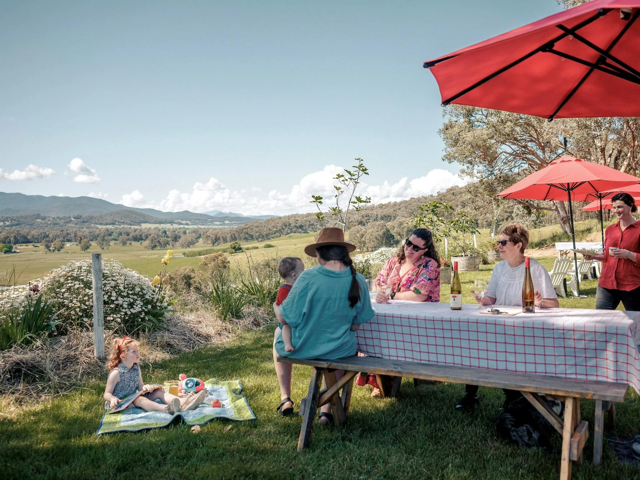 People enjoying outdoor gathering with a scenic countryside view, picnic table with wine bottles, children playing on a blanket, and red umbrellas providing shade.