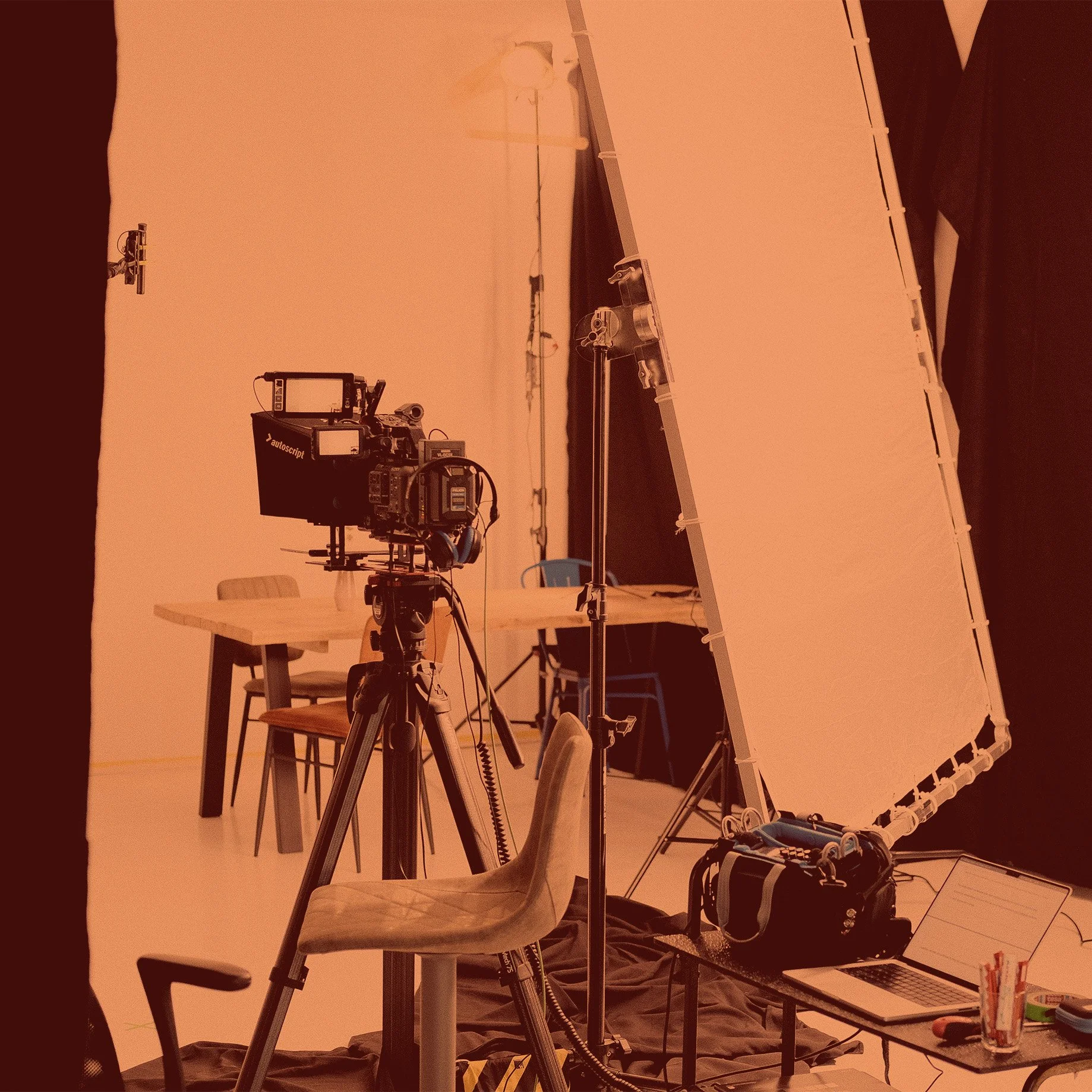Foto eines Filmstudios mit Kameraausrüstung, darunter eine Kamera auf einem Stativ, ein großer Reflexschirm, ein Tisch im Hintergrund und diverses Zubehör wie Laptop und Stühle. Die Szene hat eine orangefarbene Beleuchtung.