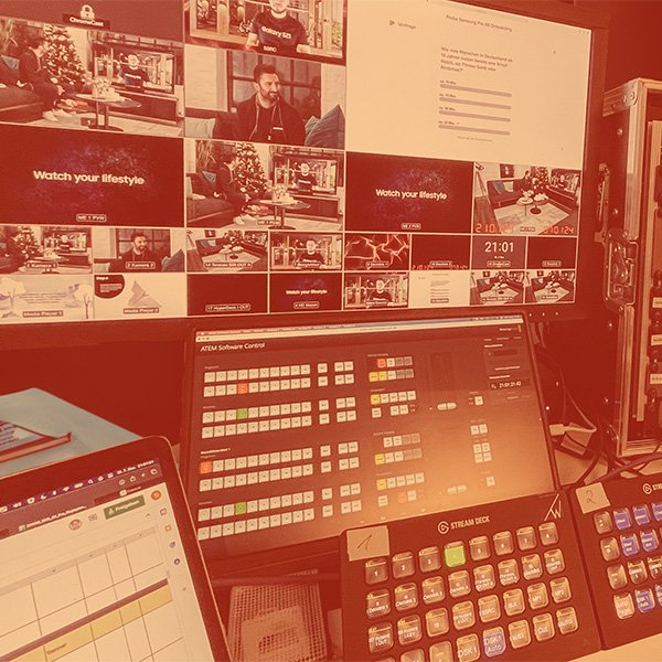Regieraum mit mehreren Bildschirmen und Steuergeräten, die TV-Produktion steuern. Überwachungsbildschirme zeigen verschiedene TV-Inhalte, während Steuerpulte wie der Stream Deck verwendet werden, um die Übertragung zu bearbeiten. Notizen und ein Laptop im Vordergrund unterstützen die Produktionsarbeit.