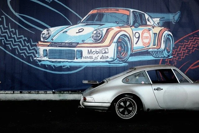 Ein silberner Porsche steht vor einem großen Wandbild, das einen stilisierten Porsche-Rennwagen mit der Nummer 9 zeigt.