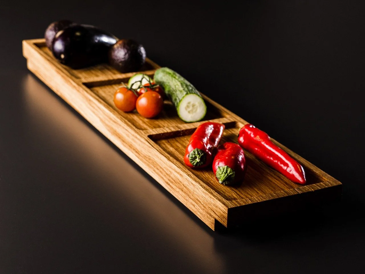 Holztablett mit Paprika, Gurke, Tomaten und Avocados auf schwarzem Hintergrund.