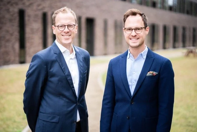 Zwei Männer in Anzügen stehen draußen vor einem Gebäude. Sie lächeln und schauen in die Kamera.