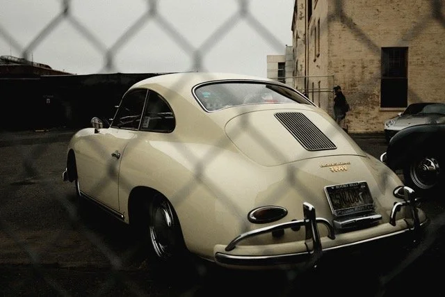 Klassisches Auto, möglicherweise ein älteres Modell, geparkt hinter einem Maschendrahtzaun. Rückansicht zeigt runde Karosserie, Chromstoßstangen und seitliche Fenster. Hintergrund mit historischen Gebäuden.