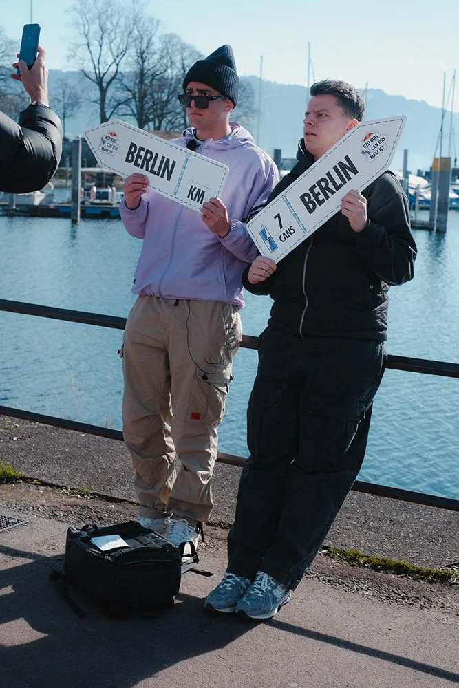 Zwei Personen stehen nebeneinander und halten Schilder mit der Aufschrift 'BERLIN' in den Händen. Einer der Schilder zeigt 'KM', während der andere '7 CANS' anzeigt. Im Hintergrund ist ein Wasser mit Booten und Bäumen zu sehen.
