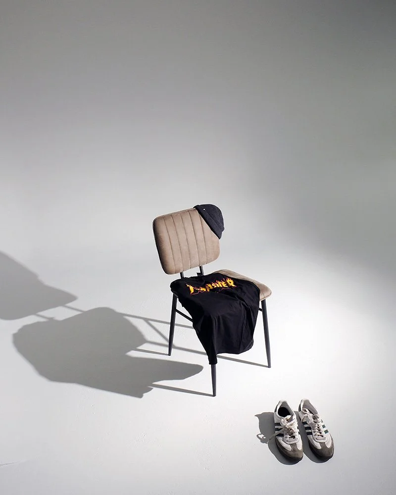 Stuhl mit schwarzem T-Shirt, beige-graue Schuhe auf dem Boden, grauer Hintergrund.