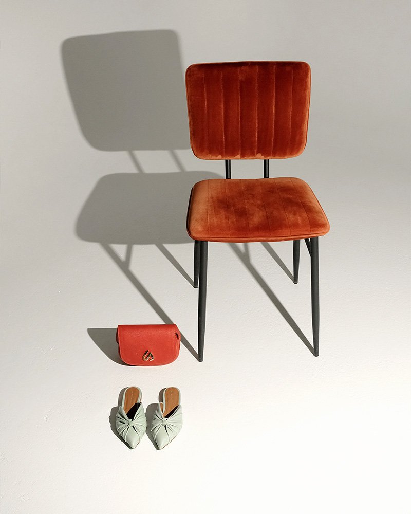 Brauner Stuhl mit Metallbeinen auf einem hellen Hintergrund. Darunter eine rote Handtasche und ein Paar helle Damenschuhe.