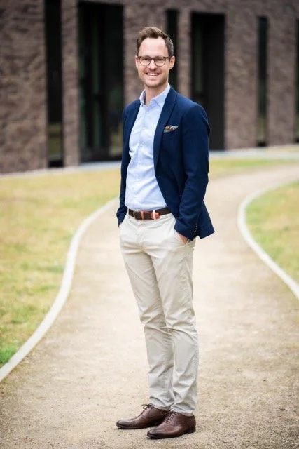 Ein lächelnder Mann in einem blauen Blazer und heller Hose steht auf einem Weg vor einem Gebäude.