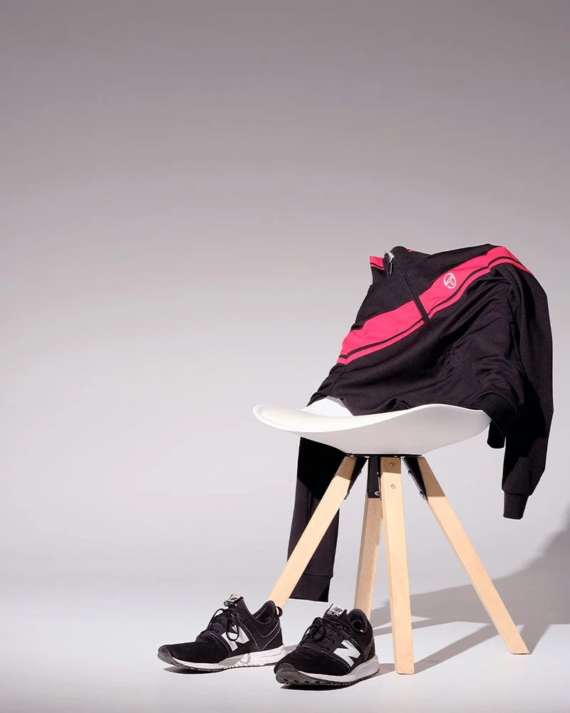 Schwarz-pinkfarbene Sportjacke auf einem weißen Stuhl mit Holzbeinen, daneben schwarze Sportschuhe auf dem Boden.