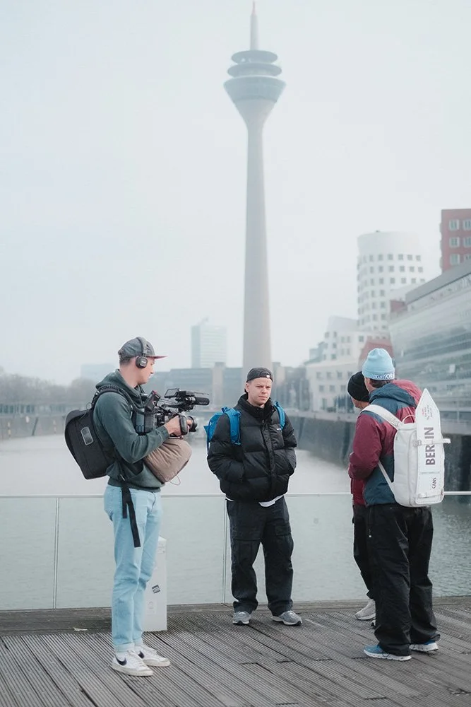 Drei Personen stehen auf einer Promenade vor dem Rheinturm in Düsseldorf. Eine Person hält eine Kamera und filmt. Im Hintergrund sind moderne Gebäude und der Fernsehturm zu sehen.