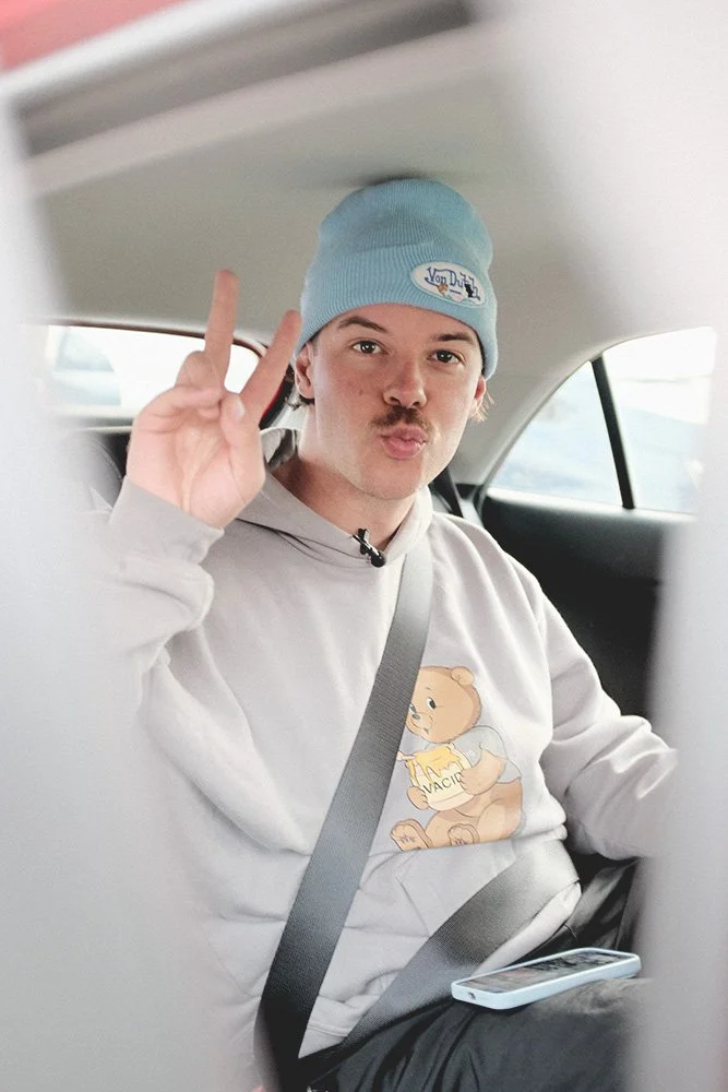 Person im Auto mit blauem Mütze und beigefarbenem Sweatshirt, zeigt Friedenszeichen, Sicherheitsgurt angelegt.