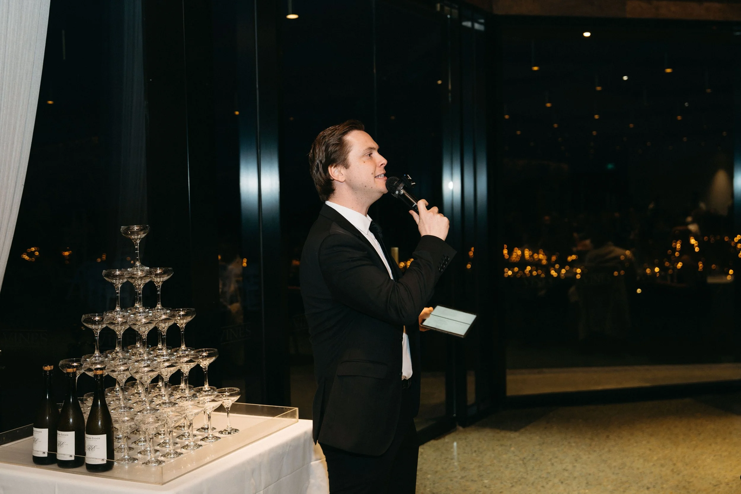 Melbourne marraige celebrant in a black suit giving a speech with a microphone at an event, next to a pyramid of champagne glasses and bottles on a table.